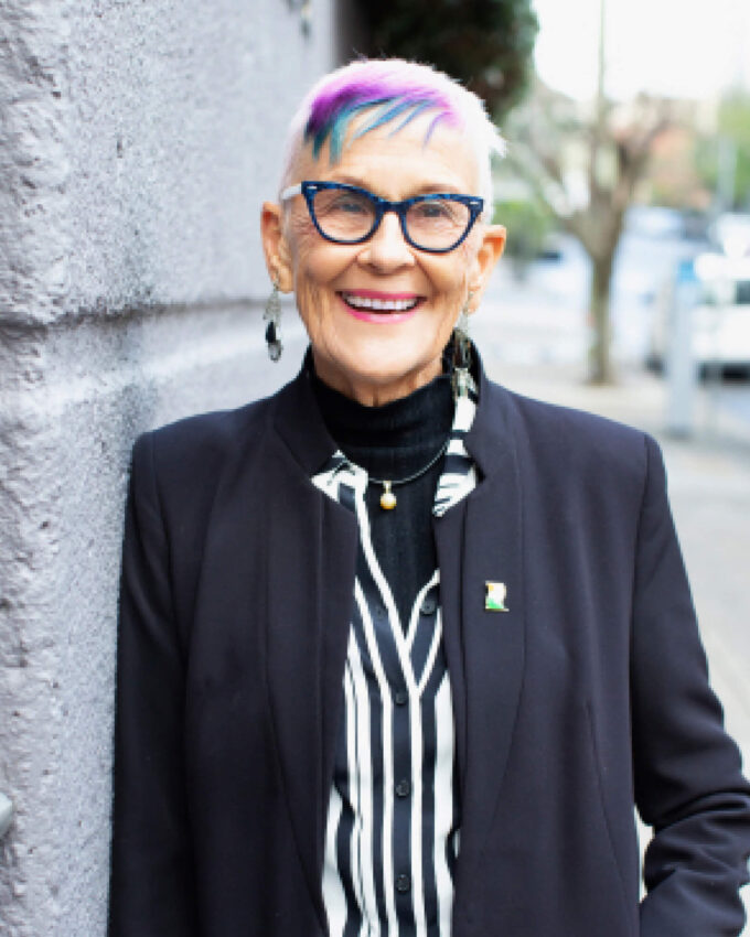 A woman with short hair coloured purple and blue is leaning against a grey wall. She is wearing a black blazer and black glasses.